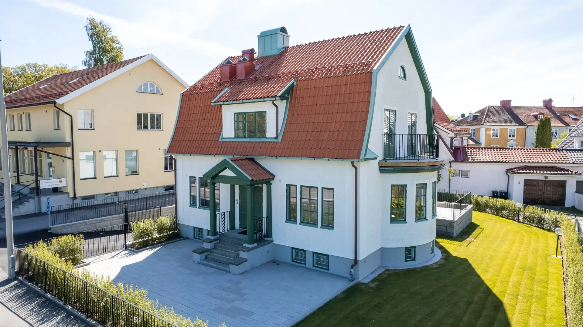 Vit villa med rött tegeltak och gröna fönsterbågar i soligt villaområde.